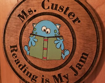 Puede incluir: Un letrero redondo de madera con un monstruo azul leyendo un libro. El letrero dice "Ms. Custer Reading is My Jam".