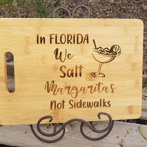 May include: A wooden cutting board with a handle, featuring the text "In Florida We Salt Margaritas Not Sidewalks" and a drawing of a margarita glass.
