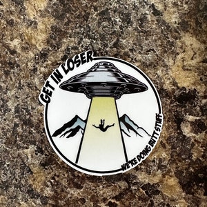 May include: A white circle sticker with a black and white illustration of a UFO with a person falling from it. The text "Get in Loser, We're Doing Butt Stuff" is written around the edge of the sticker.