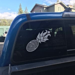 Op de afbeelding: Een witte ananassticker is bevestigd op de achterruit van een blauwe vrachtwagen. Het ananasontwerp is gestileerd met een gedetailleerd kruispatroon en een bladrijke kroon. De achtergrond toont een huis en een gedeeltelijk bewolkte lucht.