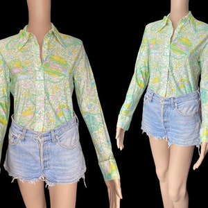 Pode incluir: Uma camisa de manga comprida com botões e um padrão floral verde, amarelo e rosa. A camisa tem gola e fechamento com botões na frente. A modelo está usando a camisa com shorts jeans.