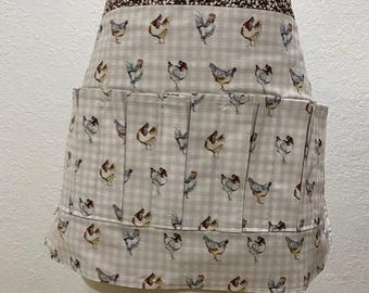 May include: A waist apron featuring a light beige and white gingham pattern with a repeating print of various chickens. The apron has a dark brown waistband with a small white floral pattern and includes multiple pleated pockets across the front.