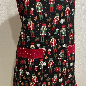 May include: A black apron featuring a repeating pattern of nutcrackers, gingerbread men, candy canes, and stockings. The apron includes two front pockets and a bottom ruffle, both in red fabric with white polka dots, and red trim along the edges.