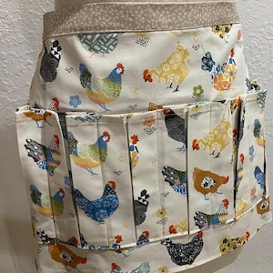 May include: A cream-colored apron with a repeating pattern of colorful chickens. The apron has multiple pockets and a pleated design. The top band and pocket trim feature a complementary floral pattern. The chickens are in various colors, including blue, yellow, and brown.