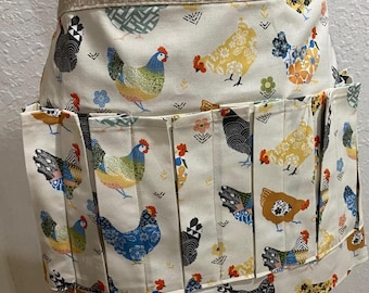May include: A cream-colored apron with a repeating pattern of colorful chickens. The apron has multiple pockets and a pleated design. The top band and pocket trim feature a complementary floral pattern. The chickens are in various colors, including blue, yellow, and brown.