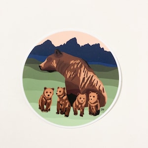 May include: A brown bear with four cubs in a circular sticker. The background is a green field with a blue mountain range in the distance. The sky is a light pink color.
