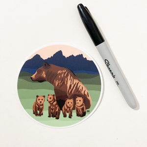 May include: A brown bear with four cubs in a mountain landscape. The bear and cubs are standing on a green grassy area with a blue lake and mountains in the background. The sky is a light pink color.