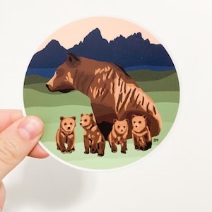 May include: A brown bear with four cubs in a mountain landscape. The bear and cubs are standing on a green field with a blue lake and mountains in the background. The sky is a light orange color.
