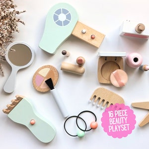 May include: A 16 piece wooden beauty playset with a hairdryer, makeup compact, comb, hairbrush, mirror, lipstick, eyeshadow palette, hair ties, and a star-shaped accessory. The playset is in pastel pink, green, and brown colors.