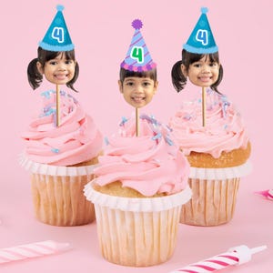 May include: Three cupcakes with pink frosting and a face cutout on a stick, each wearing a party hat with the number 4. The cupcakes sit on a pink background with candles.