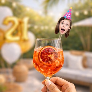 May include: A clear glass filled with an orange cocktail garnished with a blood orange slice. A custom drink stirrer features a cutout of a person's face wearing a pink party hat with blue polka dots and the number 21 printed in white on the front.