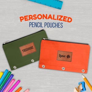 May include: Two personalized pencil pouches, one olive green and one orange, with black zippers and leather name tags. The olive green pouch has the name "THEODORE" and the orange pouch has the name "Luna". The text "PERSONALIZED PENCIL POUCHES" is at the top.