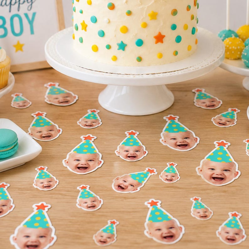 May include: A collection of paper confetti pieces featuring a smiling baby wearing a teal party hat with yellow polka dots and an orange star topper, scattered across a wooden surface near a white cake stand and a cake with matching decor.