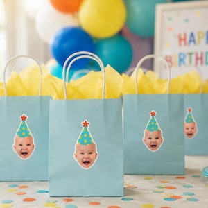 May include: Light blue paper gift bags with white twisted paper handles, each featuring a sticker of a baby wearing a teal party hat with yellow polka dots and a red star on top, filled with yellow tissue paper for a birthday celebration.