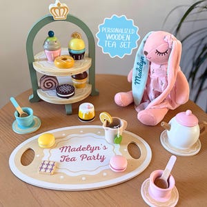 May include: A wooden tea set with a tiered dessert stand, a pink plush bunny, and a serving tray. The tray reads "Madelyn's Tea Party." The set includes a teapot, teacups, and miniature desserts. The text "Personalized Wooden Tea Set" is also visible.