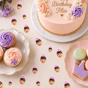 May include: Custom party confetti featuring small circular cutouts of a child's face wearing a pink crown. The confetti is scattered around a peach-colored birthday cake with the text Happy Birthday Mia, purple and peach cupcakes, and cookies.