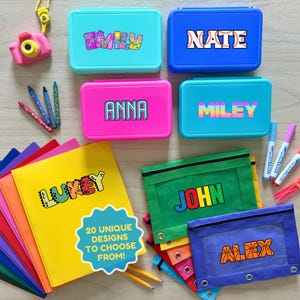 May include: A collection of colorful school supplies, including lunchboxes, pencil cases, and notebooks, each personalized with a name. The lunchboxes are in various colors like blue, pink, and teal. The image also includes crayons, markers, and a toy camera.