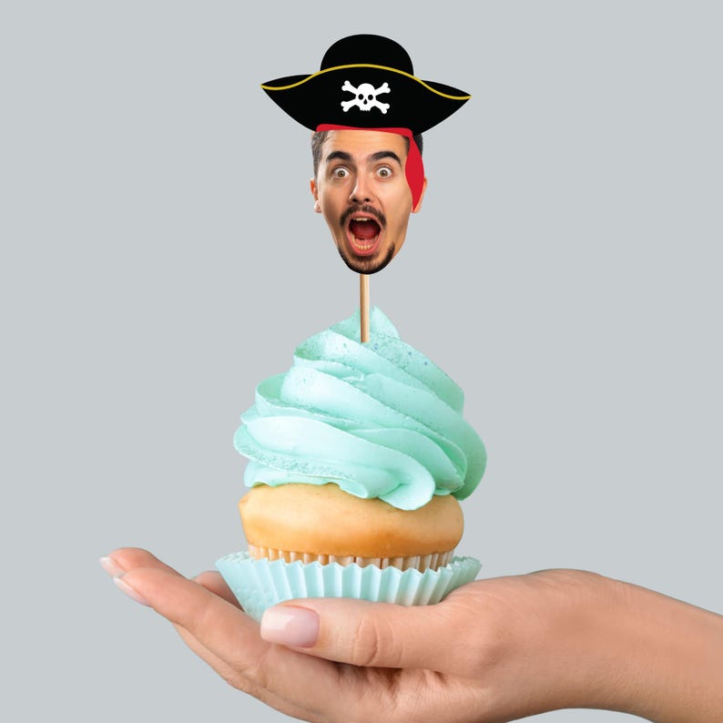 May include: A cupcake with light blue frosting and a pirate-themed topper. The topper features a person's face with a pirate hat and a red bandana. The cupcake is in a light blue paper cup.