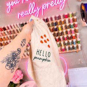 Pode incluir: Uma t-shirt de cor creme com um bolso no peito com o texto bordado "HELLO pumpkin" em fio verde escuro, acentuado por pequenos ícones de abóboras cor de laranja costurados acima do bolso no tecido.