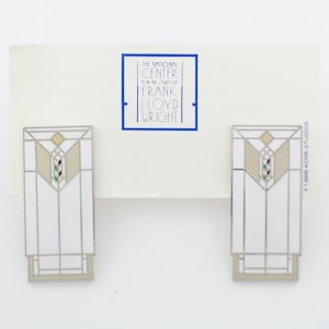 May include: A pair of white enamel earrings with a silver-toned frame, featuring a geometric design inspired by Frank Lloyd Wright's architecture. The earrings are packaged on a card with the text "The National Center for the Study of Frank Lloyd Wright" and "© 1988 Acme Studios".
