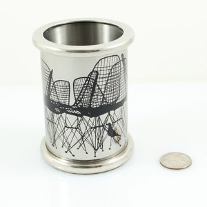 May include: A silver metal pencil holder with a black silhouette design of chairs and a bird. The design is reminiscent of a roller coaster.