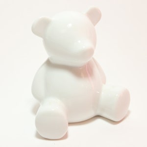 May include: A white ceramic teddy bear figurine. The bear is sitting with its legs crossed and its head tilted slightly to the side.