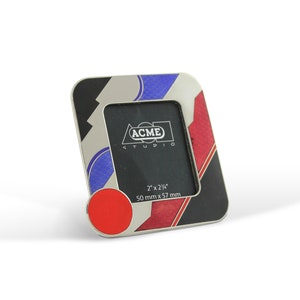 May include: A silver picture frame with a black, red, and blue geometric design. The frame is 2 inches by 2 1/4 inches and measures 50 mm by 57 mm. The frame has a clear glass front and a black backing. The frame has the text "ACME STUDIO" printed on the front.