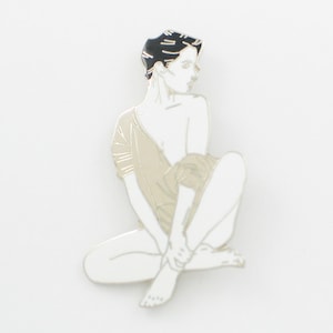May include: Enamel pin featuring a seated figure with black hair and a neutral-toned garment. The figure is rendered in white with black accents, creating a minimalist design. The pin is set against a white background.