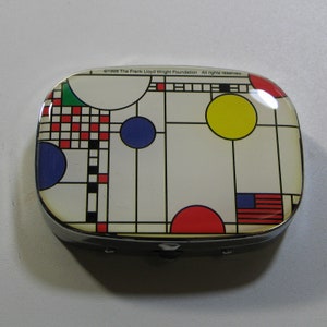 May include: A silver compact with a colorful geometric design featuring red, yellow, blue, green, and black squares and circles. The text "©1998 The Frank Lloyd Wright Foundation All rights reserved" is printed on the top of the compact.