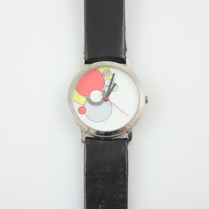 May include: A silver wristwatch with a black leather band. The watch face has a white background with a colorful abstract design of circles and a red hour hand and black minute hand.