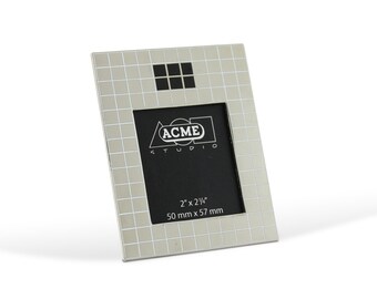 May include: Silver picture frame with a grid pattern and a black mat. The frame is 2 inches by 2 1/4 inches and measures 50 mm by 57 mm. The frame has the text "ACME STUDIO" printed on the mat.