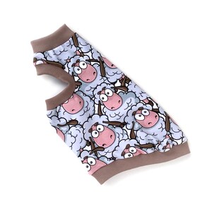 May include: A dog sweater featuring a repeating pattern of cartoon sheep with pink faces and brown horns. The sweater has a light blue background and brown trim around the neck and armholes. The garment is made of a soft material.