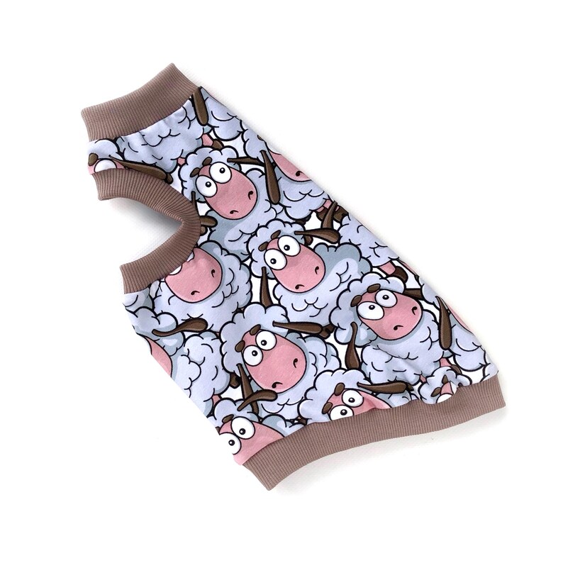 May include: A dog sweater featuring a repeating pattern of cartoon sheep with pink faces and brown horns. The sweater has a light blue background and brown trim around the neck and armholes. The garment is made of a soft material.