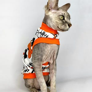 May include: A gray Sphynx cat wearing a white and orange shirt with a cartoon design. The shirt has black accents and the text "Daffy Duck" in a stylized font. The cat is sitting and looking to the right.