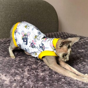 May include: A gray cat wearing a white and yellow pet shirt. The shirt has a pattern of cartoon mice and bees. The cat is stretching on a gray, textured surface.