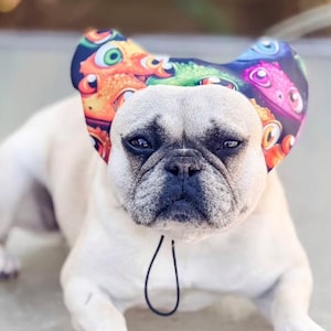 May include: A cream-coloured French Bulldog wearing a fabric headpiece with rounded ear-like flaps. The headpiece features a colourful, repeating pattern of cartoon-style lizard or dragon faces in shades of orange, green, and pink on a black background.