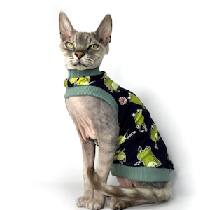 May include: A gray Sphynx cat wearing a navy blue sweater with a pattern of green frogs. The sweater has a light green collar and trim. The cat is sitting upright against a white background.