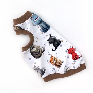 May include: White pet shirt featuring a pattern of cartoon cats in various colors and expressions. The shirt has a brown collar and trim. The cats are in shades of gray, black, orange, and red, with a watercolor effect. The background has gray and brown splatters.