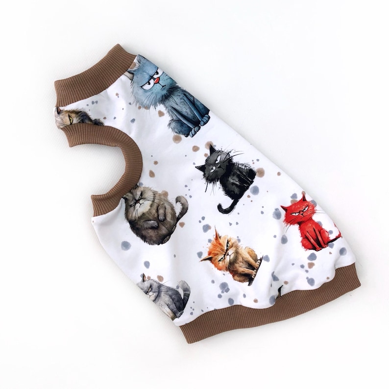 May include: White pet shirt featuring a pattern of cartoon cats in various colors and expressions. The shirt has a brown collar and trim. The cats are in shades of gray, black, orange, and red, with a watercolor effect. The background has gray and brown splatters.