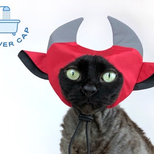 May include: A black cat wearing a red and gray shower cap with horn-like accents. The cap has a drawstring closure and the text "SHOWER CAP" is in a blue circle in the upper left corner. The cat has green eyes.