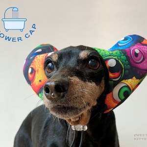 May include: A black and brown dog wearing a colourful shower cap with a multi-coloured eye pattern. The cap is made of fabric and has a shower cap design with the words "Shower Cap" printed on it.