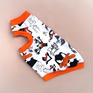 May include: White pet shirt featuring a Daffy Duck cartoon print in black and orange. The shirt has orange trim around the neck and armholes. The words "Daffy Duck" are printed on the shirt.