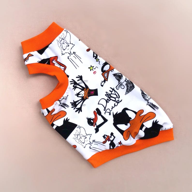 May include: White pet shirt featuring a Daffy Duck cartoon print in black and orange. The shirt has orange trim around the neck and armholes. The words "Daffy Duck" are printed on the shirt.