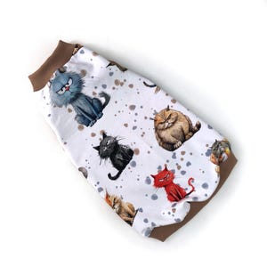 May include: White pet sweater featuring a repeating pattern of cartoon cats in various colors and expressions. The sweater has a brown ribbed collar and hem. The cats are in shades of gray, black, red, and brown, with angry or grumpy faces.