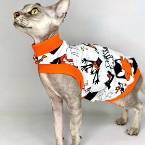 May include: A gray Sphynx cat wearing a white and orange patterned shirt. The shirt features cartoon characters in black and orange on a white background. The shirt has an orange collar and trim.