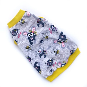 May include: A gray pet shirt with a repeating pattern of cartoon cats, mice, bees, and paw prints. The shirt has a yellow collar and sleeve cuffs. The fabric is a soft, lightweight material.