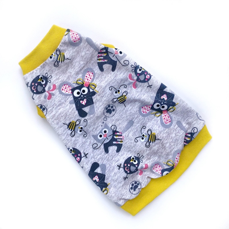May include: A gray pet shirt with a repeating pattern of cartoon cats, mice, bees, and paw prints. The shirt has a yellow collar and sleeve cuffs. The fabric is a soft, lightweight material.