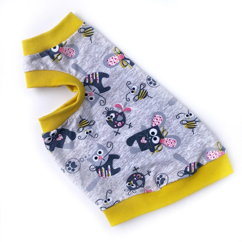 May include: Gray pet shirt with a repeating pattern of cartoon-like creatures and bees. The shirt has yellow trim around the neck and armholes. The fabric is a soft, lightweight material.