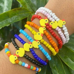 Puede incluir: Una colección de pulseras de cuentas coloridas. Cada pulsera presenta un esquema de color diferente, incluyendo arcoíris, negro, azul, amarillo, naranja, rojo, rosa y blanco. Cada pulsera tiene un dije de pato amarillo.