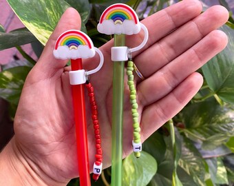 Puede incluir: Dos pajitas reutilizables con adornos de arcoíris y nubes. Una pajita es roja con un dije de cuentas rojas y la letra "P". La otra pajita es verde con un dije de cuentas verdes y la letra "T".